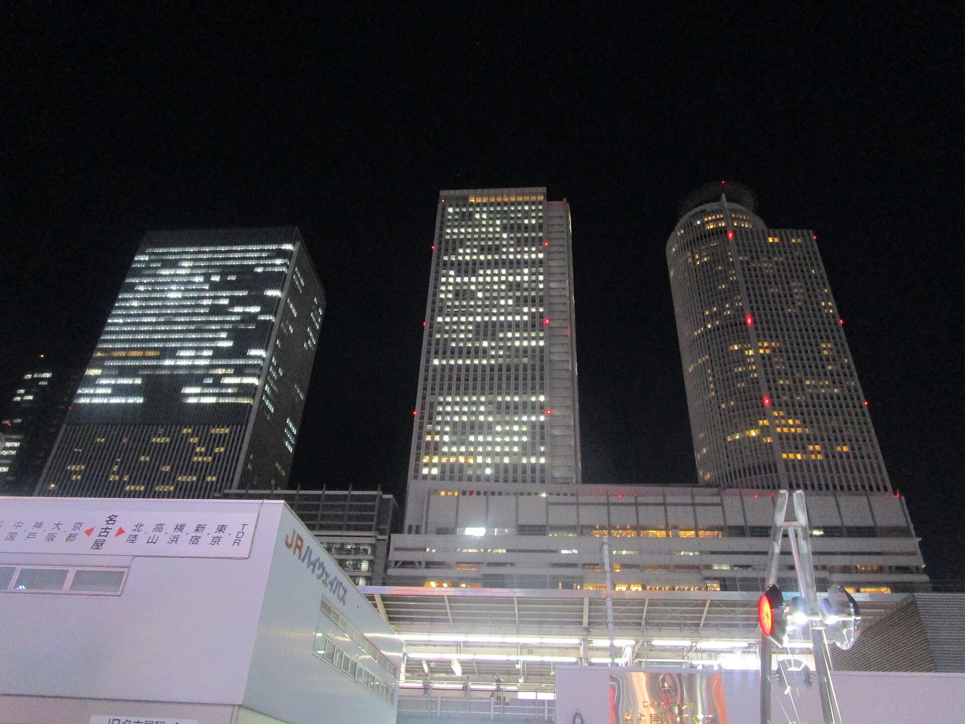 The illuminated JR Central Towers and JR Gate Tower at Nagoya Station at night, with a highway bus sign reading "Nagoya" in the foreground.