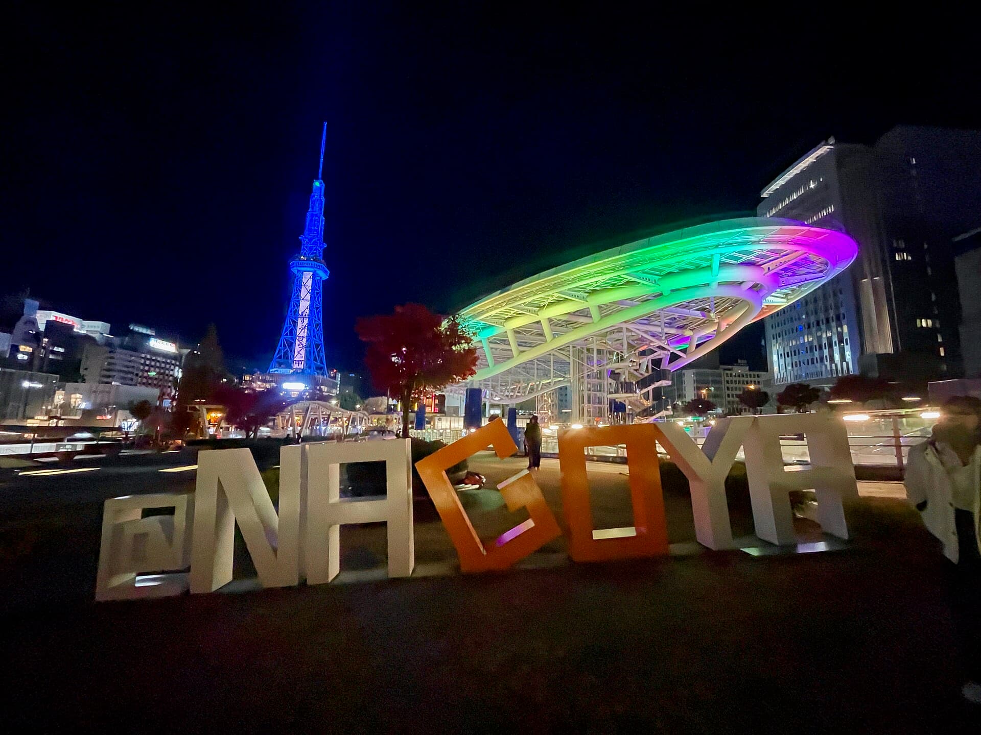 Oasis 21's illuminated glass structure and the blue-lit Nagoya TV Tower (MIRAI Tower) at night, with an "@NAGOYA" sign in the foreground.
