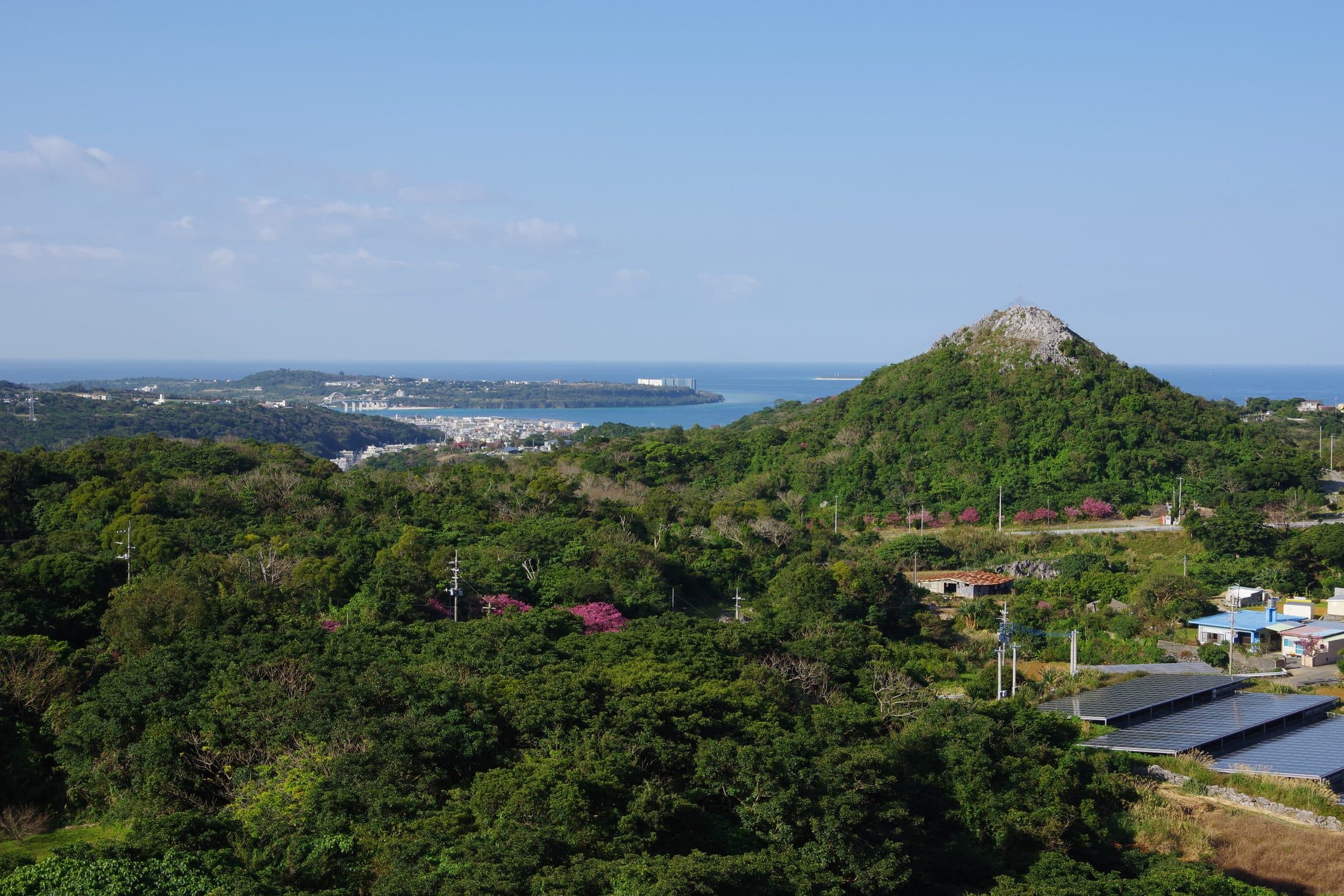 An elevated view of a bay and hillside village on the Motobu Peninsula, with houses and greenery in the foreground.