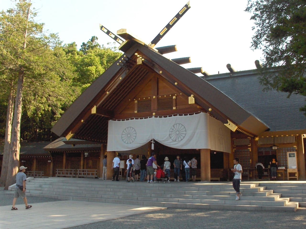 The main worship hall of Hokkaido Jingu in Maruyama, Sapporo, with visitors gathered at the entrance and forest trees surrounding the plaza.