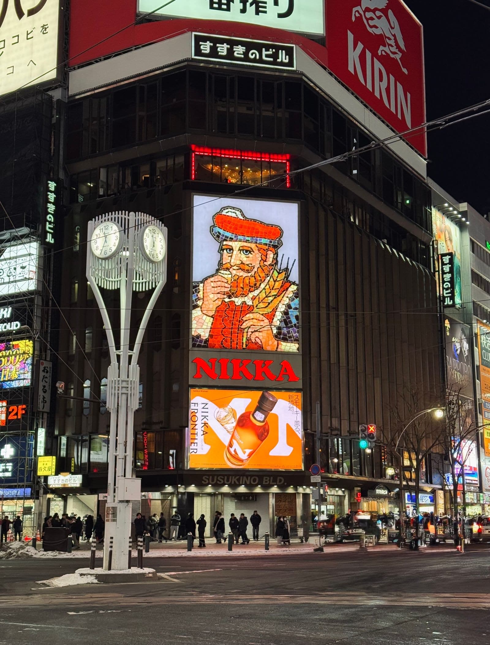 The illuminated Nikka whisky sign and clock tower at the Susukino intersection in Sapporo at night, surrounded by neon storefronts and streetlights.