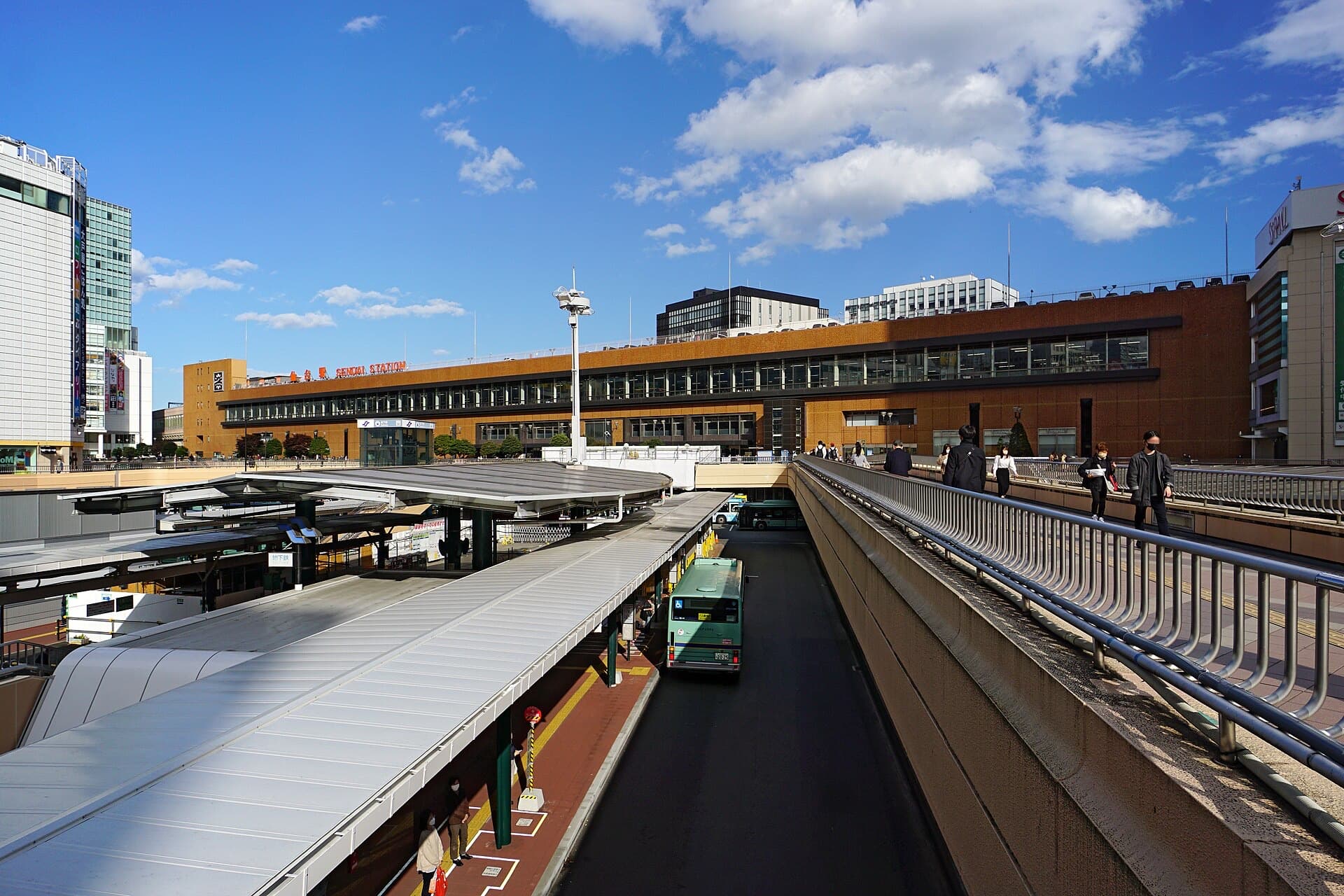 The exterior of Sendai Station with its bus terminal, elevated pedestrian deck, and "SENDAI STATION" signage under a blue sky.