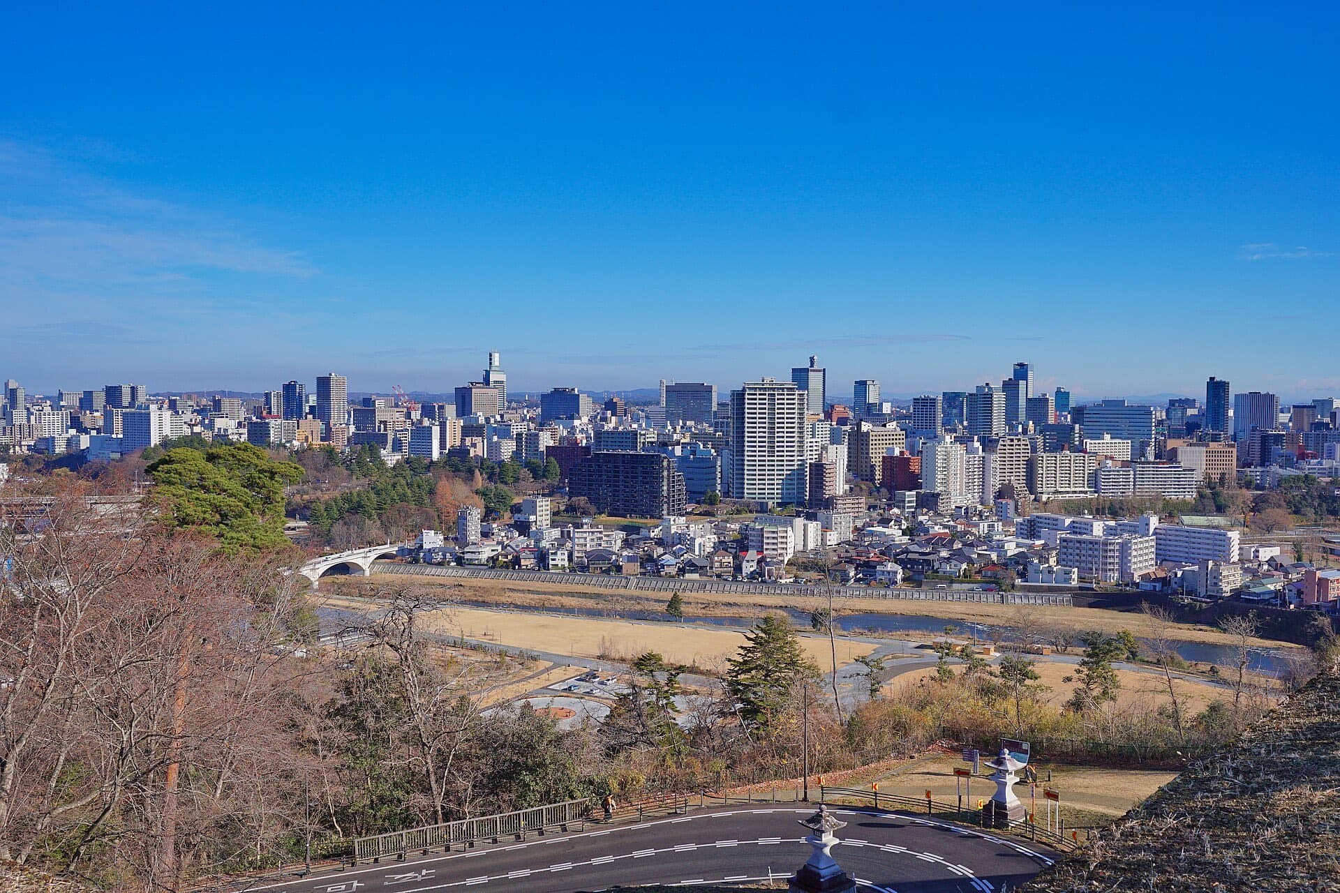 The Sendai city skyline seen from the Sendai Castle ruins, with the Hirose River, a bridge, and foreground trees under a clear blue sky.