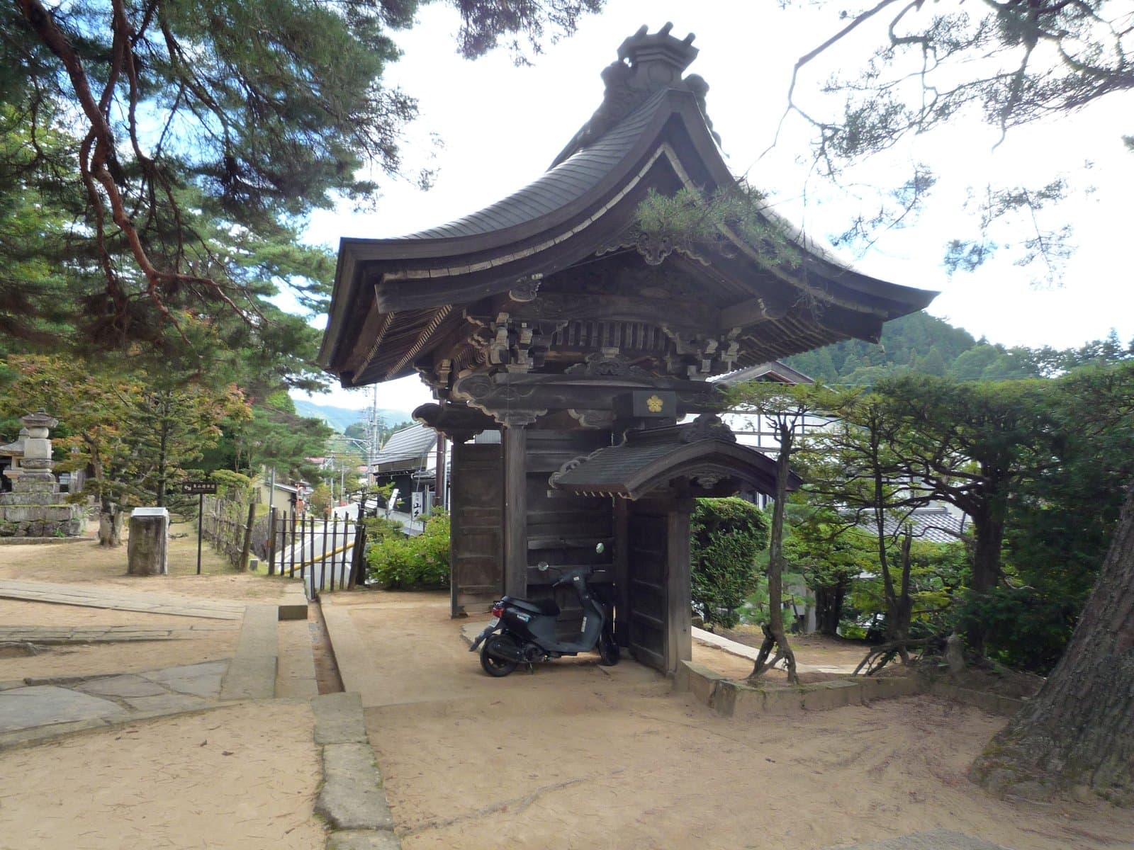 A traditional wooden temple gate along the Higashiyama Walking Course in Takayama, surrounded by pine trees with hills visible beyond.