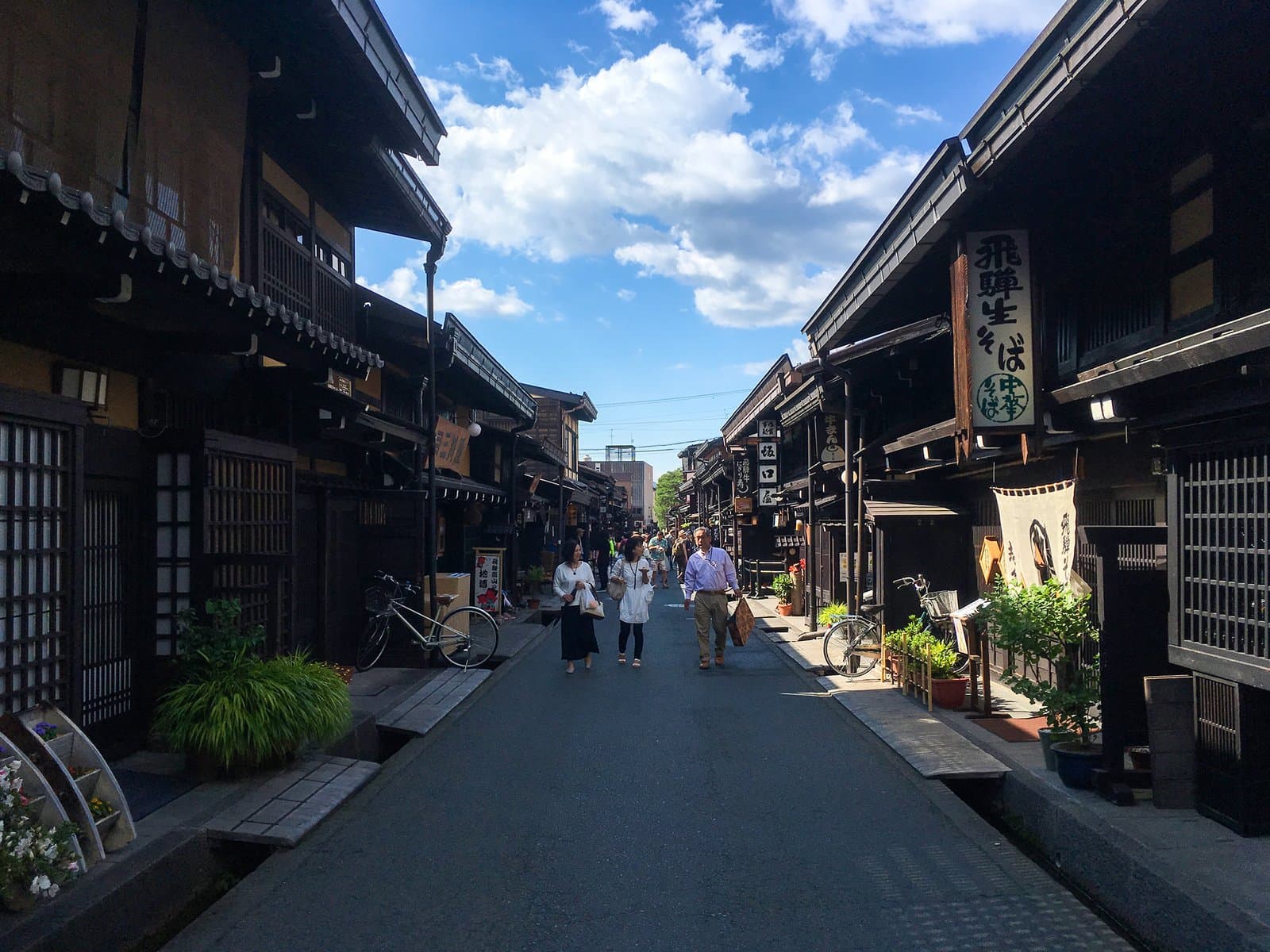 A preserved street in Takayama's Sanmachi Suji district lined with dark wooden merchant buildings and shop signs, with visitors walking beneath a partly cloudy sky.