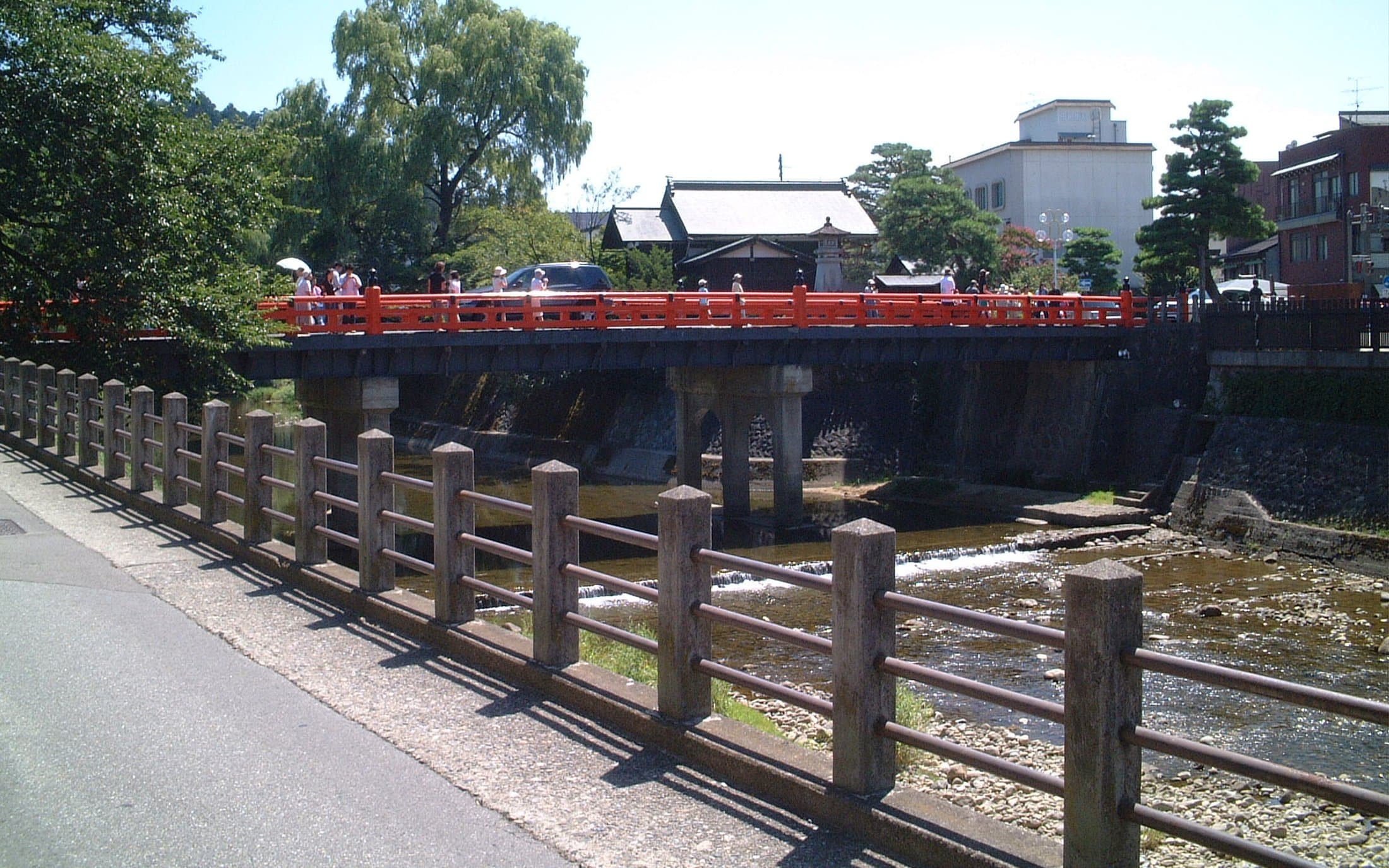 The vermillion Nakabashi Bridge crosses the Miyagawa River in Takayama, with trees, a traditional wooden building, and the town visible on both banks under a clear sky.